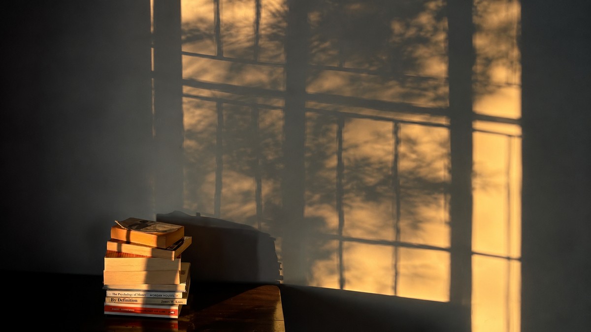 Hourglass casting a long shadow beside a stack of books, representing the slow, invisible work of long-term transformation